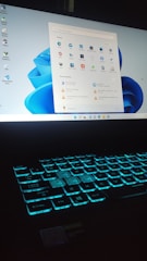 A laptop with a glowing backlit keyboard and a desktop screen displaying various icons and applications. The keyboard emits a blue-green light, creating a futuristic look. The desktop screen shows a start menu with pinned and recommended applications, set against a light blue background.