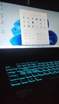 A laptop with a glowing backlit keyboard and a desktop screen displaying various icons and applications. The keyboard emits a blue-green light, creating a futuristic look. The desktop screen shows a start menu with pinned and recommended applications, set against a light blue background.