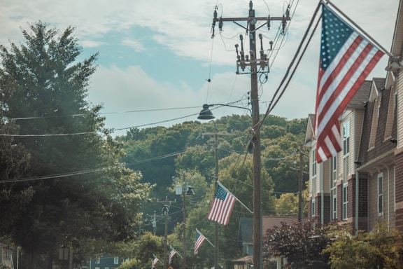 A street lined with American flags hanging on poles and attached to houses. The scene includes utility poles with cables and a backdrop of lush green hills under a partly cloudy sky.