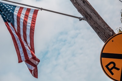 An American flag is suspended from a wooden pole against a backdrop of a partially cloudy sky. To the right of the image, there is a yellow circular sign with a black line and an 'R' partially visible.