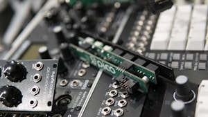 A close-up of electronic audio equipment featuring several modular synthesizer components, including circuit boards and knobs with various labels. The details highlight connectors, switches, and various controls for sound modulation.