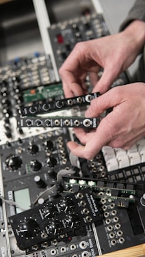 Hands are adjusting black electronic modules with various knobs and ports. The modules are part of a more extensive setup of electronic equipment, possibly a modular synthesizer, featuring various buttons and inputs.