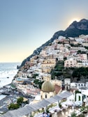 An Italian coastal village nestled in the hillside, featuring a myriad of colorful buildings stacked one over the other. The scene includes a number of small boats dotting the calm, azure sea. In the foreground, the dome of a church stands out with its intricate design, while vibrant greenery surrounds the area. The setting sun casts a gentle glow over the landscape.