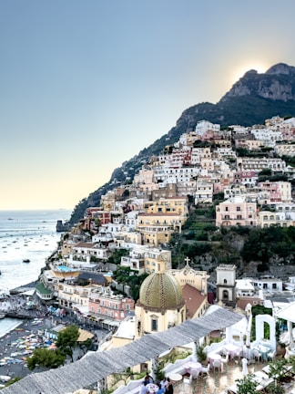 An Italian coastal village nestled in the hillside, featuring a myriad of colorful buildings stacked one over the other. The scene includes a number of small boats dotting the calm, azure sea. In the foreground, the dome of a church stands out with its intricate design, while vibrant greenery surrounds the area. The setting sun casts a gentle glow over the landscape.