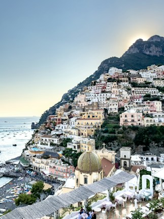 An Italian coastal village nestled in the hillside, featuring a myriad of colorful buildings stacked one over the other. The scene includes a number of small boats dotting the calm, azure sea. In the foreground, the dome of a church stands out with its intricate design, while vibrant greenery surrounds the area. The setting sun casts a gentle glow over the landscape.