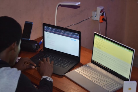 A person is seated at a wooden desk working with two laptops. One laptop displays code or a technical document, while the other shows a web interface page. Additional items on the desk include a smartphone, a lamp, and multiple power outlets. The setting appears to be a workspace focused on programming or technical tasks.