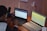 A person is seated at a wooden desk working with two laptops. One laptop displays code or a technical document, while the other shows a web interface page. Additional items on the desk include a smartphone, a lamp, and multiple power outlets. The setting appears to be a workspace focused on programming or technical tasks.