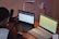 A person is seated at a wooden desk working with two laptops. One laptop displays code or a technical document, while the other shows a web interface page. Additional items on the desk include a smartphone, a lamp, and multiple power outlets. The setting appears to be a workspace focused on programming or technical tasks.