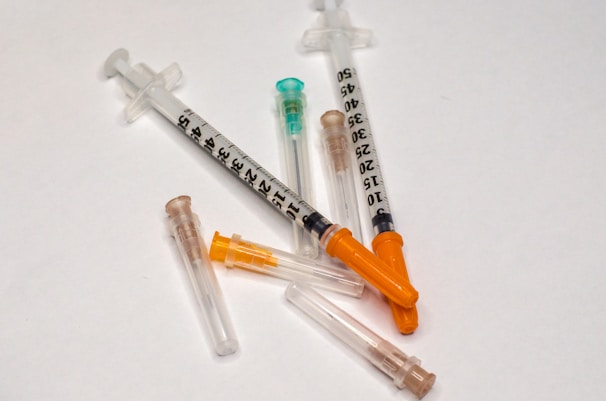 Several syringes and needles scattered on a white surface. The syringes have orange and green caps, with black numerical markings visible on the barrels.