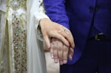 A couple is holding hands, with the focus on their hands. The woman's hand is adorned with intricate henna designs and two rings, while the man's hand has a simple ring. They are dressed in formal attire, with the woman's outfit featuring ornate beadwork and embroidery, and the man's outfit being a blue suit.