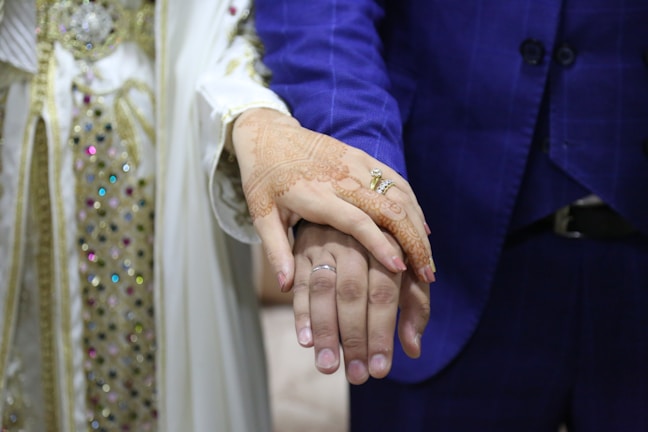 A couple is holding hands, with the focus on their hands. The woman's hand is adorned with intricate henna designs and two rings, while the man's hand has a simple ring. They are dressed in formal attire, with the woman's outfit featuring ornate beadwork and embroidery, and the man's outfit being a blue suit.