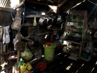 A rustic, cluttered kitchen area within a shed-like structure. The space holds multiple hanging pots and pans on the walls. There are several cleaning supplies and household items like detergent containers, strainers, and baskets scattered around. A green plastic bucket with a red cover takes center stage on the floor. To the right, there is a green-racked shelf holding various kitchen utensils and items. The area appears dark and worn but functional.