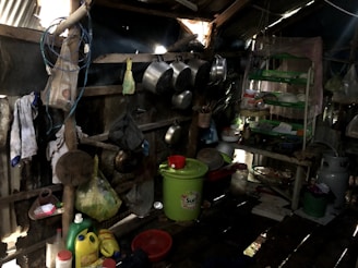 A rustic, cluttered kitchen area within a shed-like structure. The space holds multiple hanging pots and pans on the walls. There are several cleaning supplies and household items like detergent containers, strainers, and baskets scattered around. A green plastic bucket with a red cover takes center stage on the floor. To the right, there is a green-racked shelf holding various kitchen utensils and items. The area appears dark and worn but functional.