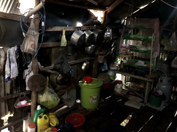 A rustic, cluttered kitchen area within a shed-like structure. The space holds multiple hanging pots and pans on the walls. There are several cleaning supplies and household items like detergent containers, strainers, and baskets scattered around. A green plastic bucket with a red cover takes center stage on the floor. To the right, there is a green-racked shelf holding various kitchen utensils and items. The area appears dark and worn but functional.