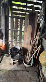 A rustic setting with wooden slats as a partial wall, showing pieces of wood leaning against the structure. There is a black, pot-like object sitting on top of a makeshift cooking stove made of blocks. Nearby are various kitchen utensils, including a basket, a small bowl, and long wooden sticks. The background shows greenery through the gaps in the wall.