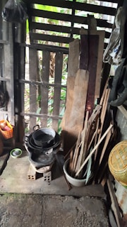 A rustic setting with wooden slats as a partial wall, showing pieces of wood leaning against the structure. There is a black, pot-like object sitting on top of a makeshift cooking stove made of blocks. Nearby are various kitchen utensils, including a basket, a small bowl, and long wooden sticks. The background shows greenery through the gaps in the wall.