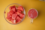 A clear bowl filled with diced watermelon pieces is positioned next to a glass mug filled with a red smoothie. Both items are placed on a vibrant yellow surface.