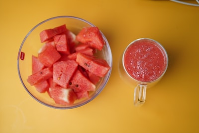 A clear bowl filled with diced watermelon pieces is positioned next to a glass mug filled with a red smoothie. Both items are placed on a vibrant yellow surface.