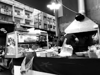 A street vendor operates a food stall on an urban street at night, with a glass display case filled with various food items. The vendor is facing away, preparing food with cooking utensils. The background shows residential buildings and electrical wires.