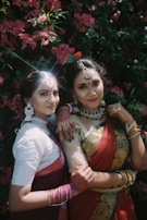 Two women dressed in traditional attire with intricate jewelry and henna designs on their hands pose in front of lush pink flowers. Their colorful sarees feature elaborate patterns.