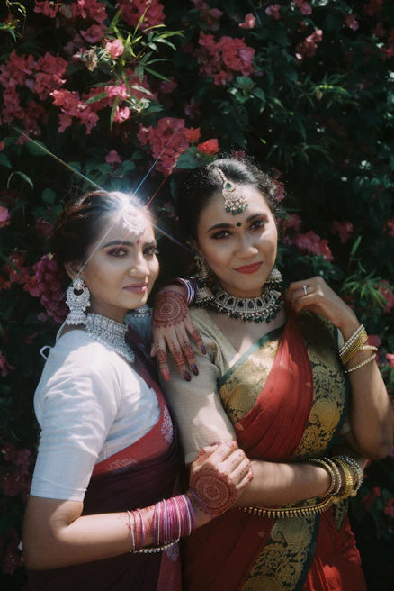 Two sisters holding hands, dressed in cream and beige kurta sets, standing in a sunlit garden with marigold decorations.