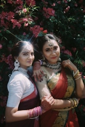 Two women dressed in traditional attire with intricate jewelry and henna designs on their hands pose in front of lush pink flowers. Their colorful sarees feature elaborate patterns.