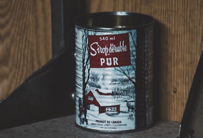 A tin can of pure maple syrup with a capacity of 540 ml is displayed. The label features a winter scene depicting a snow-covered landscape with bare trees and a red cabin. The text is written in French, emphasizing the product's authenticity. The background consists of wooden and metallic surfaces.