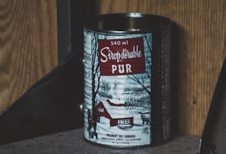 A tin can of pure maple syrup with a capacity of 540 ml is displayed. The label features a winter scene depicting a snow-covered landscape with bare trees and a red cabin. The text is written in French, emphasizing the product's authenticity. The background consists of wooden and metallic surfaces.