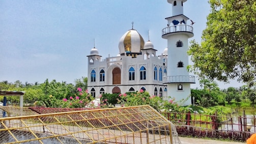A beautiful mosque with a golden dome and tall minaret surrounded by lush greenery and vibrant flowers. The building features arched windows and decorative elements, set against a clear blue sky. A fence with a net in the foreground and a tranquil pond are also visible.