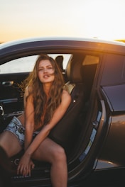 A confident female driver smiling warmly while waiting in a sleek, pink-accented car at sunset.