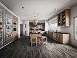 A modern kitchen with sleek wooden cabinetry and stainless steel appliances. The room features a center island with stools, a hanging light fixture, and ample natural light from large windows. Shelving displays various food and beverage items.