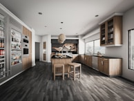 A modern kitchen with sleek wooden cabinetry and stainless steel appliances. The room features a center island with stools, a hanging light fixture, and ample natural light from large windows. Shelving displays various food and beverage items.