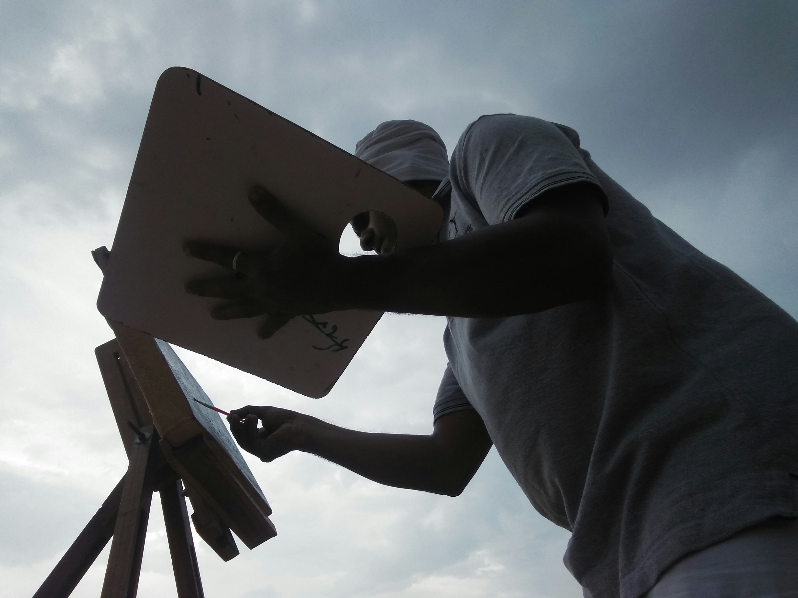 A person is silhouetted against a cloudy sky, standing in front of an easel and palette, appearing to paint or draw. The scene captures the artist in action, with a focus on the creative process.