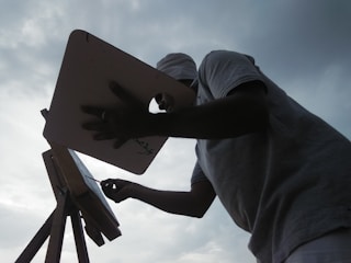 A person is silhouetted against a cloudy sky, standing in front of an easel and palette, appearing to paint or draw. The scene captures the artist in action, with a focus on the creative process.