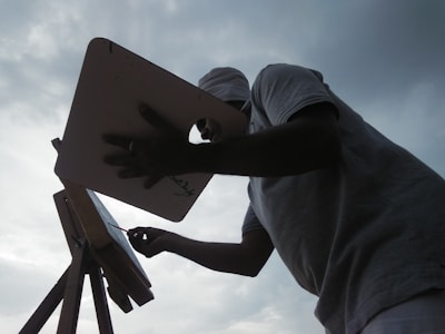 A person is silhouetted against a cloudy sky, standing in front of an easel and palette, appearing to paint or draw. The scene captures the artist in action, with a focus on the creative process.