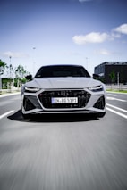 A sleek, modern car is moving at speed down a city road. The vehicle's design is aerodynamic with sharply defined lines and LED headlights. The background shows a clear sky and some urban elements like trees and a building.