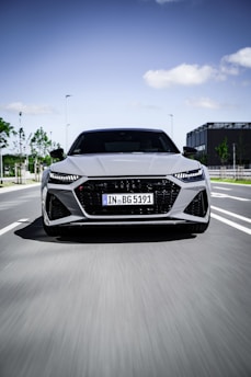 A sleek, modern car is moving at speed down a city road. The vehicle's design is aerodynamic with sharply defined lines and LED headlights. The background shows a clear sky and some urban elements like trees and a building.