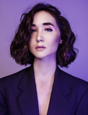 A portrait of a person with short, wavy hair and a contemplative expression, wearing a dark blazer against a purple background. The lighting emphasizes the smoothness of their skin and the subtle makeup with pink tones on the eyelids and glossy lips.