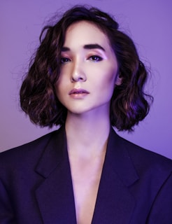 A portrait of a person with short, wavy hair and a contemplative expression, wearing a dark blazer against a purple background. The lighting emphasizes the smoothness of their skin and the subtle makeup with pink tones on the eyelids and glossy lips.