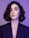 A portrait of a person with short, wavy hair and a contemplative expression, wearing a dark blazer against a purple background. The lighting emphasizes the smoothness of their skin and the subtle makeup with pink tones on the eyelids and glossy lips.