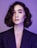A portrait of a person with short, wavy hair and a contemplative expression, wearing a dark blazer against a purple background. The lighting emphasizes the smoothness of their skin and the subtle makeup with pink tones on the eyelids and glossy lips.