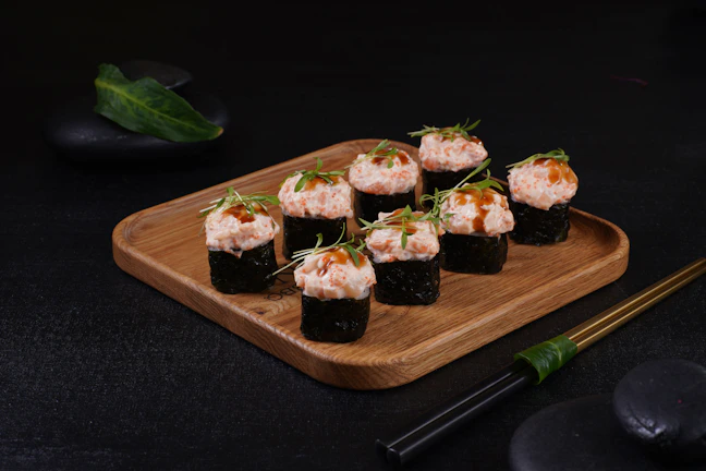 A wooden platter holds a neatly arranged set of sushi rolls topped with creamy seafood and garnished with thin green herbs. The sushi is placed in two parallel rows on the dark surface, with decorative chopsticks nearby. Smooth black stones and a green leaf add to the aesthetic presentation.