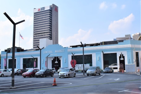 A light blue building with the words 'Central Market' on it is visible. There are several parked cars in front, and a tall building with the name 'Agro Bank' in the background. A Malaysian flag is flying on a pole, and the sky is partly cloudy.