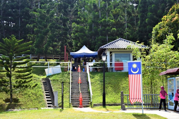 A lush outdoor setting with a small building featuring a tiled roof surrounded by dense, tall trees and greenery. There is a staircase leading up to the building, flanked by orange traffic cones. A large Malaysian flag is displayed prominently on the right, and two people wearing pink can be seen walking nearby. A tent is set up near the building, and a lamp post stands in front.