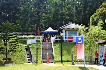 A lush outdoor setting with a small building featuring a tiled roof surrounded by dense, tall trees and greenery. There is a staircase leading up to the building, flanked by orange traffic cones. A large Malaysian flag is displayed prominently on the right, and two people wearing pink can be seen walking nearby. A tent is set up near the building, and a lamp post stands in front.