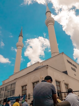 A mosque with two tall minarets rises against a bright blue sky filled with fluffy white clouds. Several people are gathered outside the building, some seated, suggesting a communal or religious gathering. The architecture is characterized by intricate details typical of Islamic design.