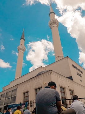 A mosque with two tall minarets rises against a bright blue sky filled with fluffy white clouds. Several people are gathered outside the building, some seated, suggesting a communal or religious gathering. The architecture is characterized by intricate details typical of Islamic design.