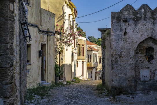 A narrow cobblestone street lined with old, rustic buildings featuring textured walls and wooden shutters. Bougainvillea vines with pink flowers climb up one of the buildings, adding a touch of color. Overhead wires stretch across the alleyway, and the warm sunlight creates soft shadows on the structures.