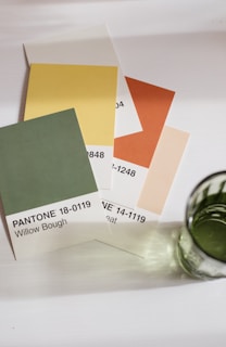 A collection of Pantone color sample cards is fanned out on a white surface. The colors displayed include green, yellow, peach, and orange, each labeled with their respective Pantone name and number. Adjacent to the cards is a green glass, casting a soft shadow on the surface.