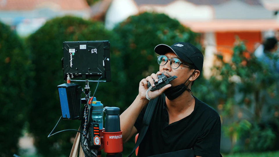 In the image, a man is standing on a street corner with his back to the camera. He's wearing a black baseball cap and glasses, and he has a drink in one hand while holding a microphone in the other. The background features an orange building and lush green trees, creating a vibrant urban setting for this scene.